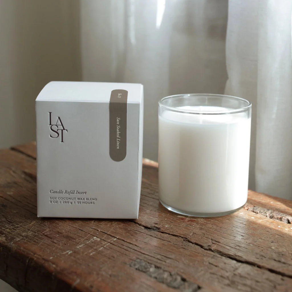 A white candle next to its packaging box on a wooden surface, with a sheer curtain in the background.