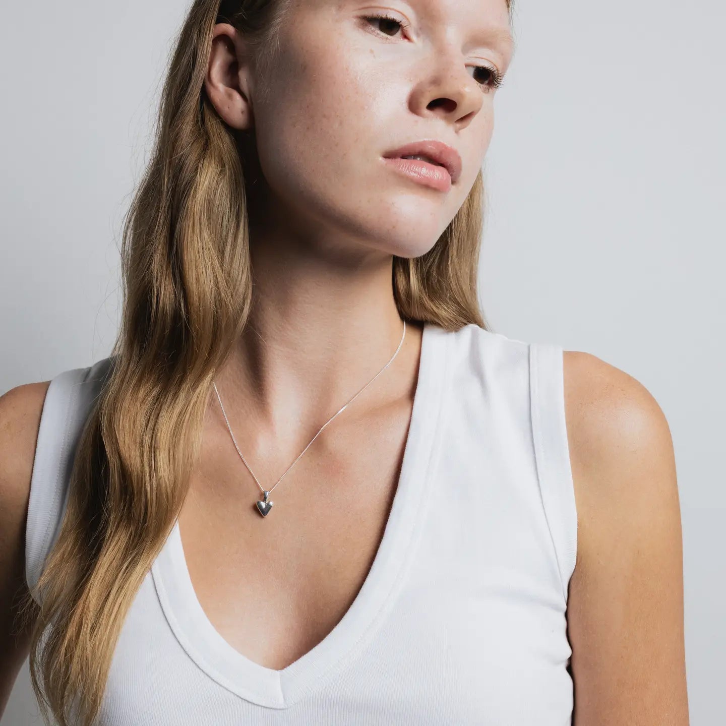 Woman wearing a white tank top and a necklace with a heart pendant on a plain background