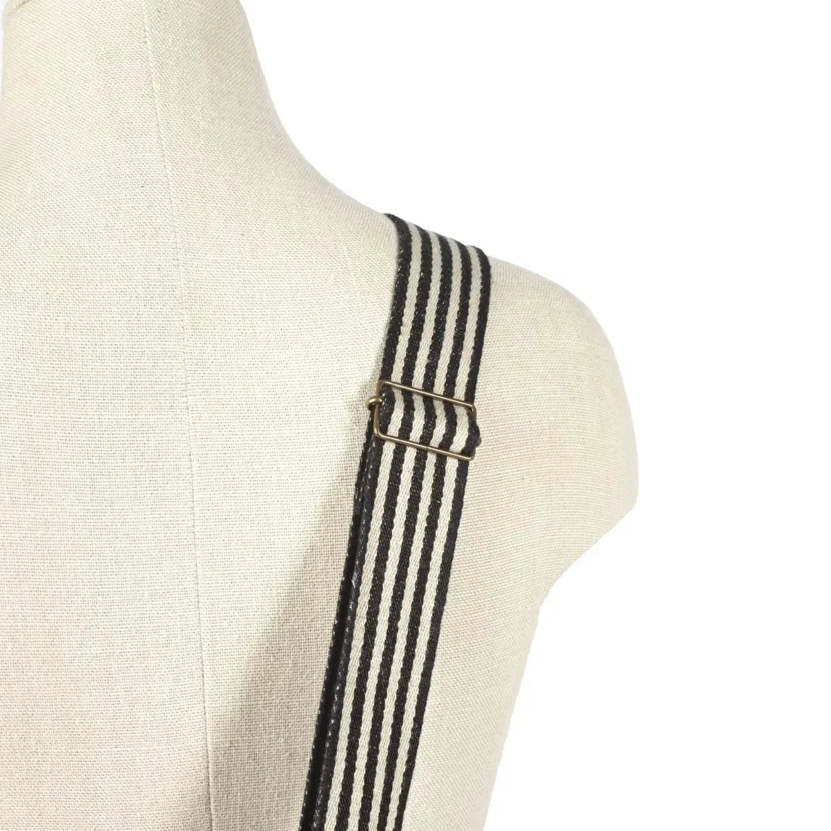 Black and white striped canvas shoulder strap with a brass buckle, shown on a cream linen mannequin torso against a white background.