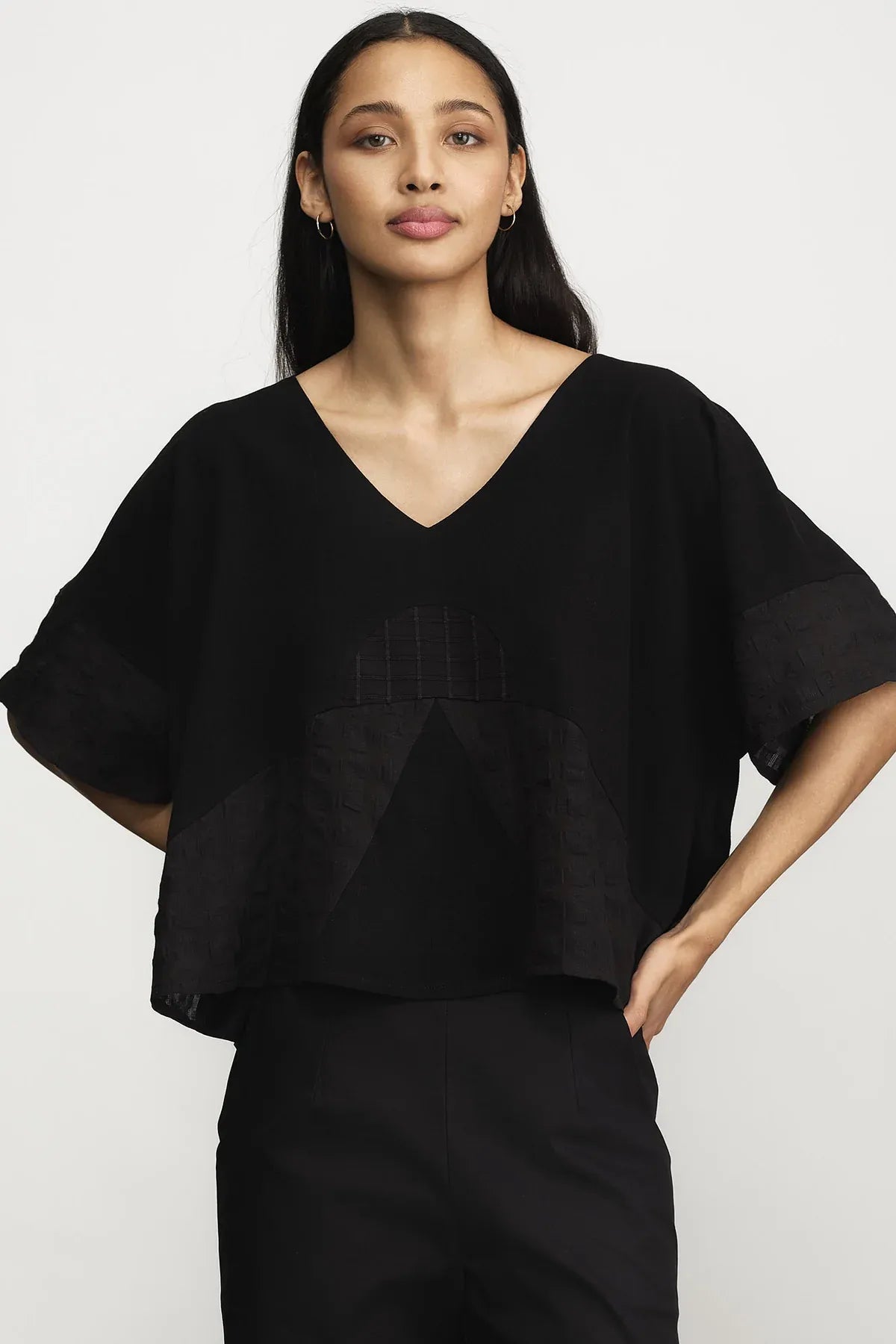 A woman modeling a black short-sleeve V-neck top with textured geometric paneling on the front, paired with black pants, against a plain light gray background.