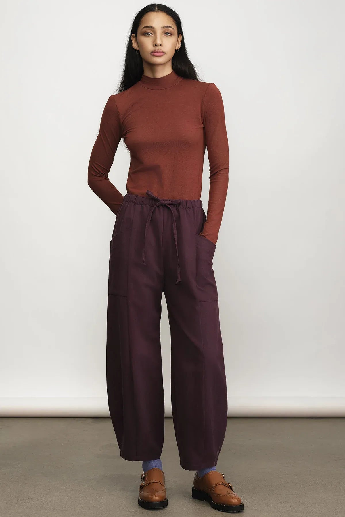 A woman modeling a rust-brown mock neck long-sleeve top and dark plum wide-leg drawstring pants, paired with brown leather loafers, against a neutral studio backdrop.