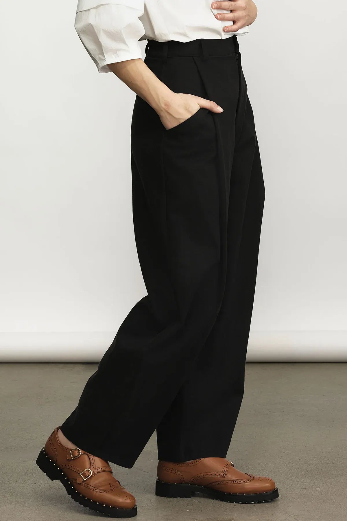 A person wearing black wide-leg trousers with a white long-sleeve top and brown leather buckle shoes, shown from the waist down against a plain studio background.