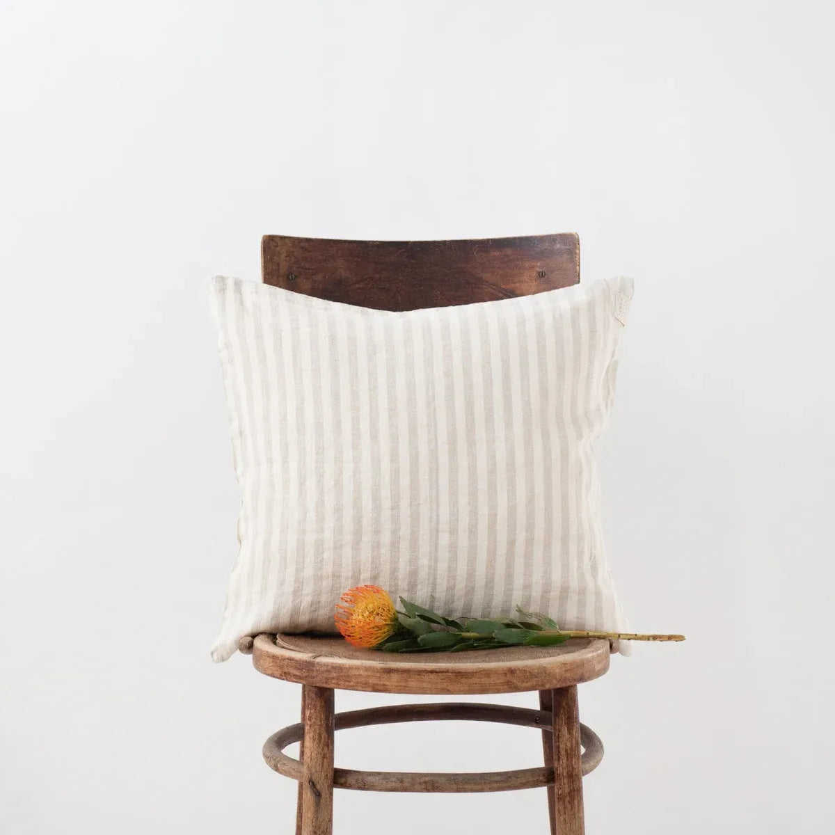 A square off-white linen throw pillow with thin vertical stripes, styled on a rustic wooden stool with a single orange flower resting on its seat, against a plain white background.
