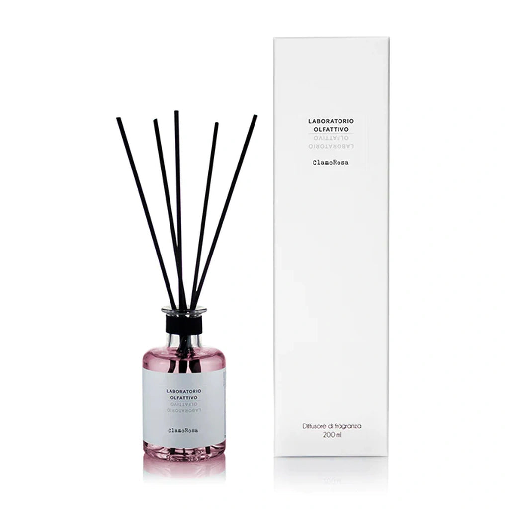 A reed diffuser bottle with pink liquid and black reeds stands next to its tall white packaging box on a clean white background
