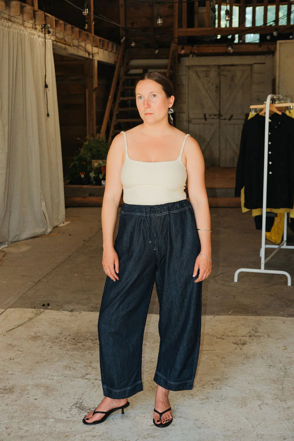 Woman wearing a white tank top and dark blue pants in an indoor setting with a wooden floor and draped fabric.
