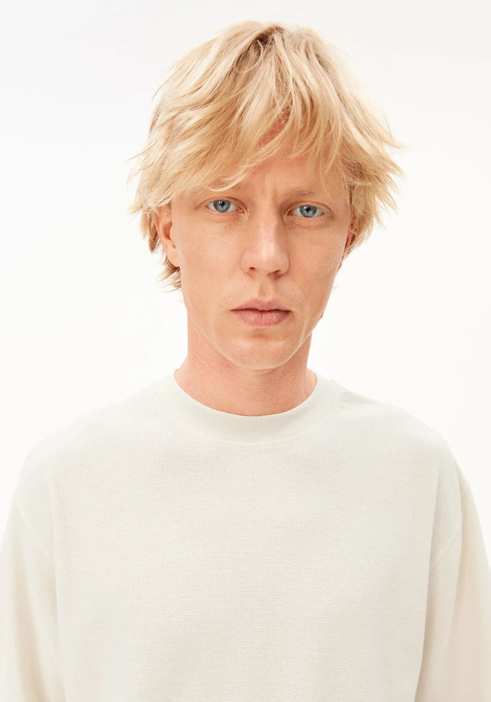 A close-up portrait of a person with short blonde hair and blue eyes, wearing a plain off-white crewneck sweater against a clean white background.