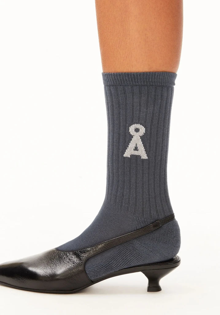 A close-up of a ribbed navy crew sock worn on a foot, featuring a white knit monogram logo, paired with a black pointed-toe kitten heel shoe against a white background.