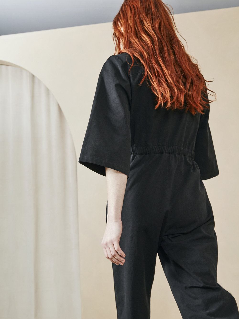 A model with long, wavy red hair is shown from behind wearing a black short-sleeve jumpsuit in a minimalist studio setting with a cream-colored background and arch detail.