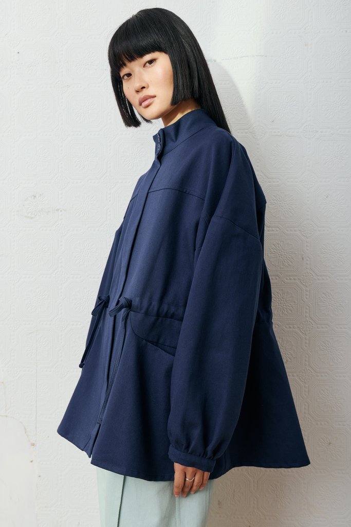 A model with a black bob haircut wearing an oversized navy blue jacket with a drawstring waist, standing against a textured off-white wall.