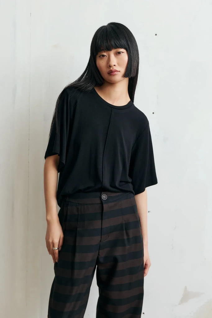 A model with a straight black haircut stands against a light off-white wall, wearing a black short-sleeve top and dark brown and black horizontally striped trousers.