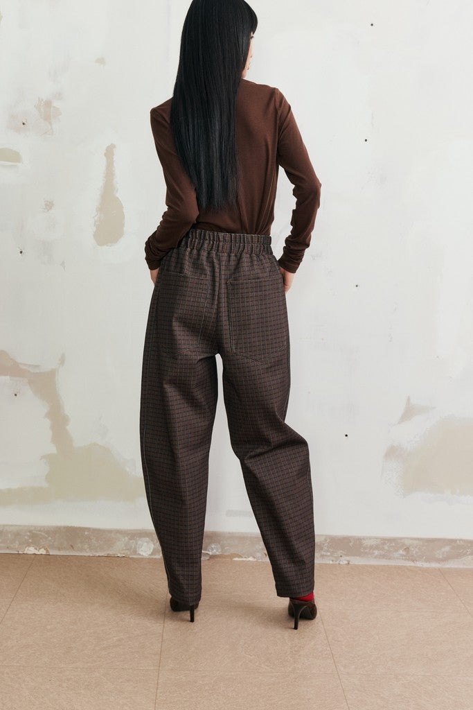A woman viewed from behind wearing a brown long-sleeve top and dark brown wide-leg trousers with a subtle small-scale check pattern, standing against a textured off-white wall.