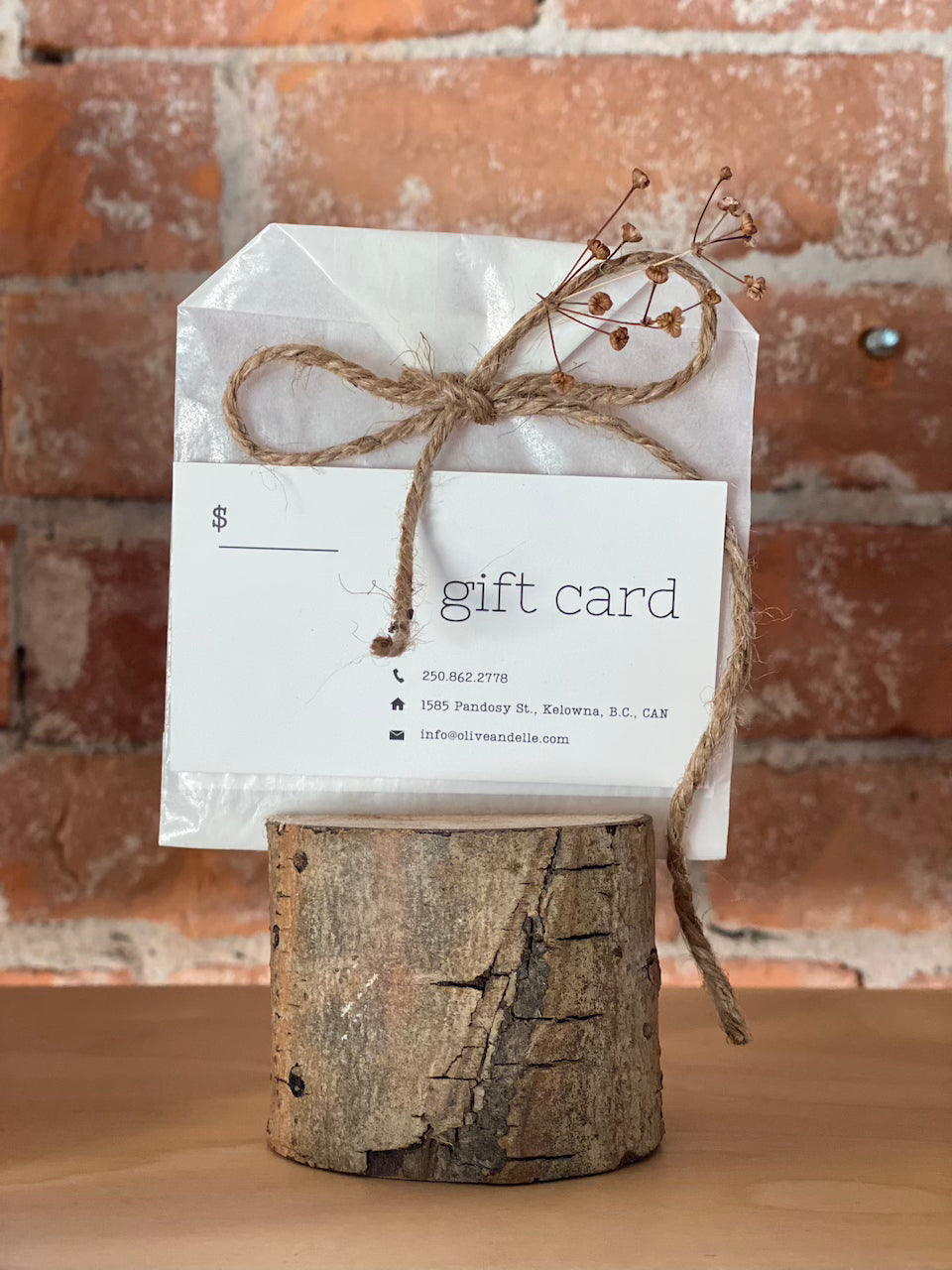 A physical gift card with 'gift card' text visible, tied with a string and presented on a wooden block with a brick wall in the background.
