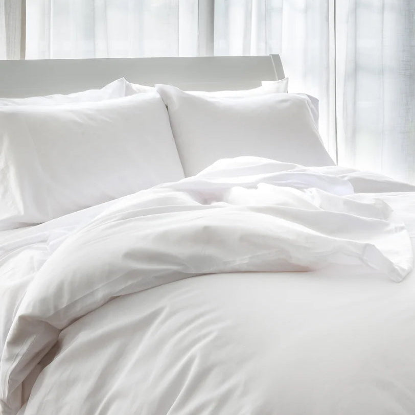 A white duvet cover with a silky appearance, displayed on a bed with pillows, in a room with a window behind.