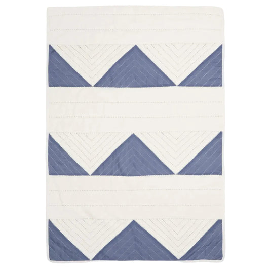 A rectangular quilted blanket featuring a geometric pattern of blue triangles and white chevron lines on an off-white background, shown flat against a light gray studio backdrop.