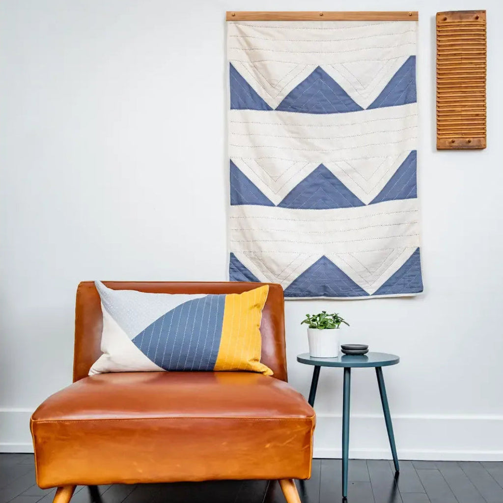 A mid-century modern tan leather armchair with wooden legs, styled with a geometric blue, white, and yellow throw pillow. A small dark teal side table with a potted plant sits next to it, all set against a white wall with a blue and white geometric wall hanging.