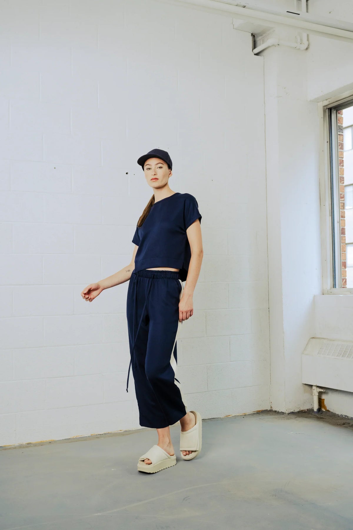 Person wearing a navy outfit standing in a minimalistic room with white walls and a window.