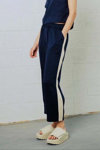 A person wearing navy cropped pants with a wide off-white side stripe, paired with cream slide sandals, standing against a white painted brick wall.