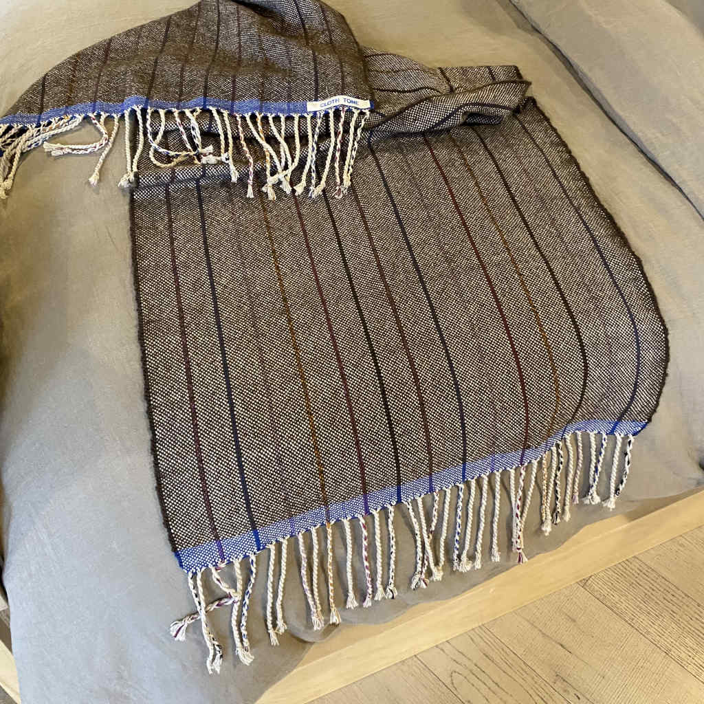 A handwoven alpaca throw in walnut color with blue fringes, laid out on a bed.