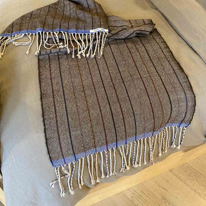 A handwoven alpaca throw in walnut color with blue fringes, laid out on a bed.