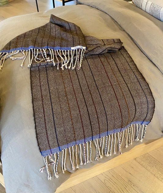 A handwoven alpaca throw in walnut color with blue fringes, laid out on a bed.