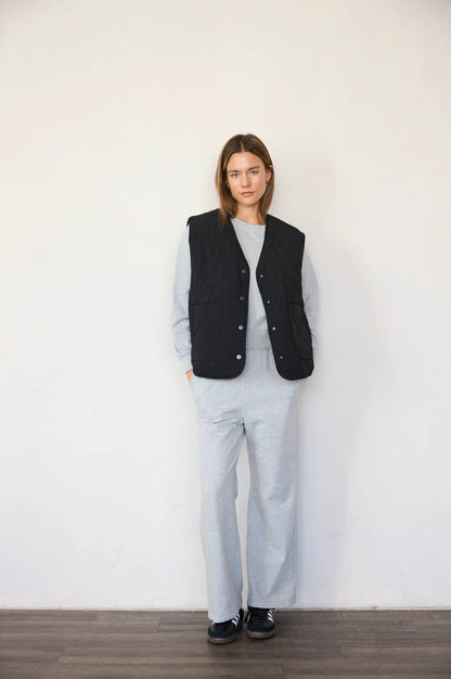 A woman stands against a plain white wall wearing a black quilted vest over a light gray long-sleeve top and matching light gray wide-leg pants, with dark slip-on shoes.