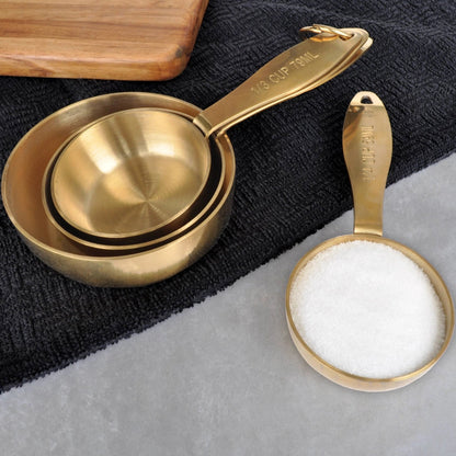 A set of gold-colored stainless steel measuring cups, including a 1/4 cup, 1/3 cup, 1/2 cup, and 1 cup size, displayed with a removable ring.