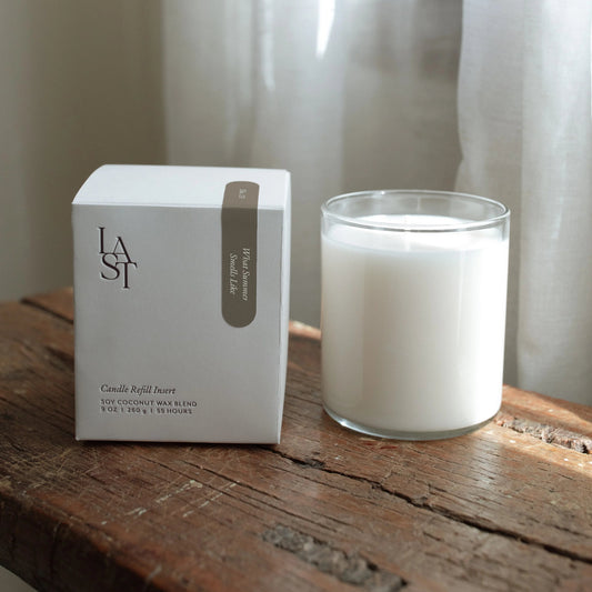 A white candle next to its packaging box on a wooden surface, with a gray and white design on the box, and a window in the background.