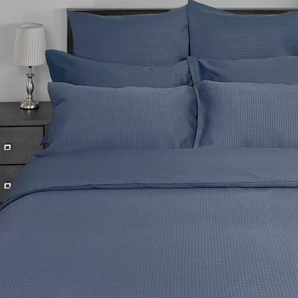 Blue textured bedding set on a bed with a nightstand and lamp in the background.
