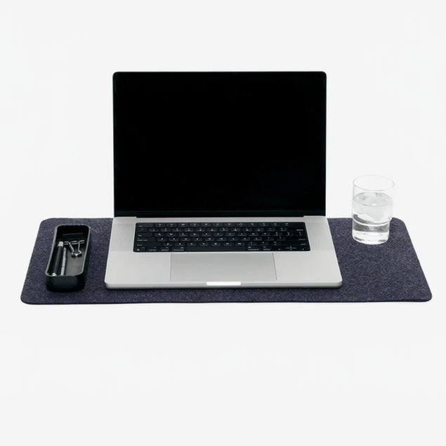 A medium-sized, charcoal-colored Merino wool desk pad displayed under a laptop and a glass, indicating its use as a protective surface for the computer and a drink.