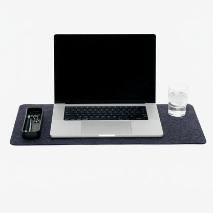A medium-sized, charcoal-colored Merino wool desk pad displayed under a laptop and a glass, indicating its use as a protective surface for the computer and a drink.