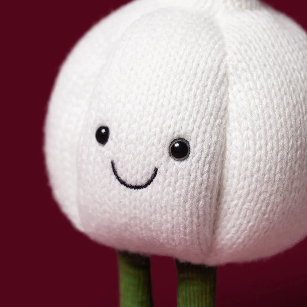 A close-up of a knitted garlic character toy with a white textured body, embroidered smiling face, and green stem-like legs against a dark red background.