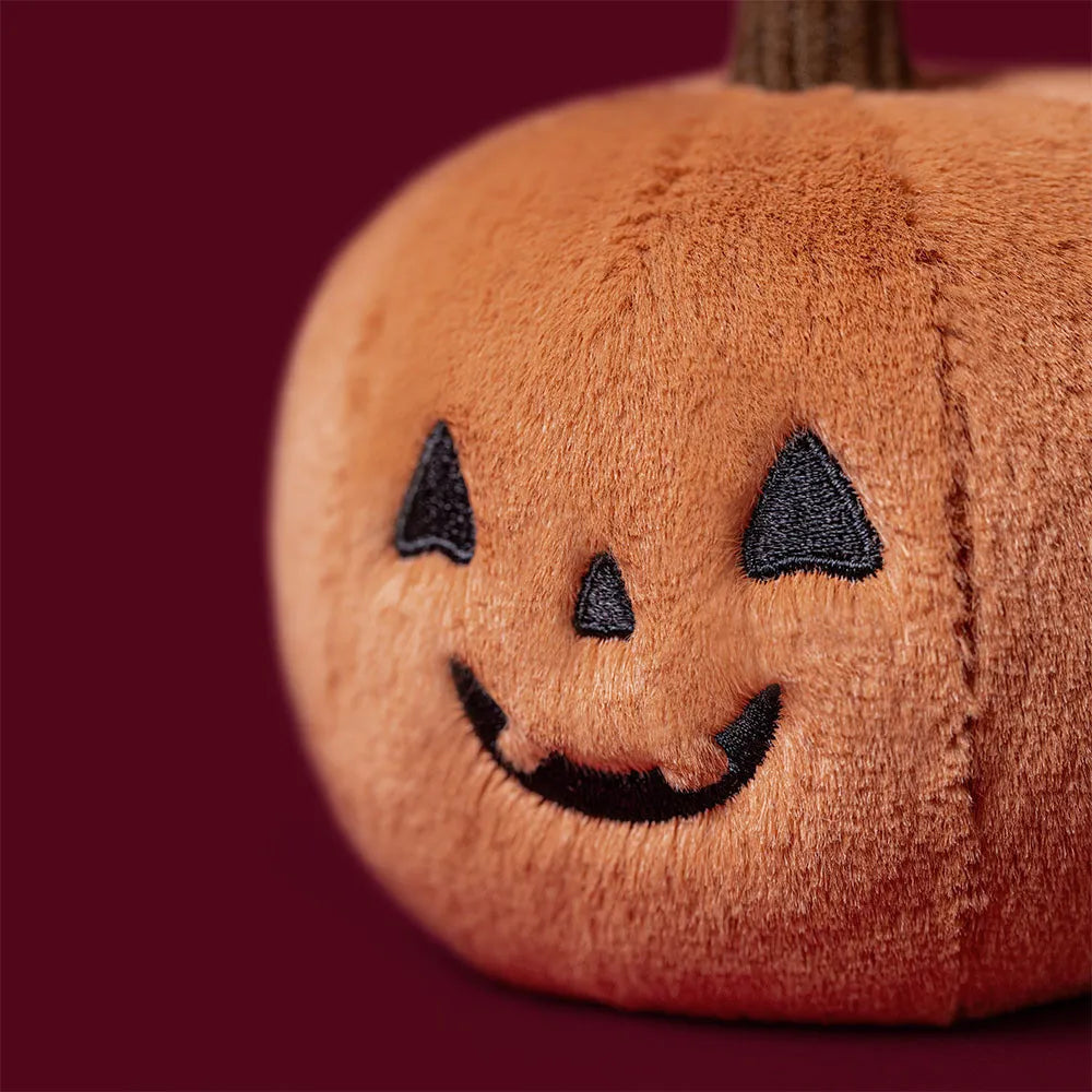 A close-up of a plush jack-o'-lantern toy with black embroidered eyes, nose, and smile, set against a solid dark burgundy background.