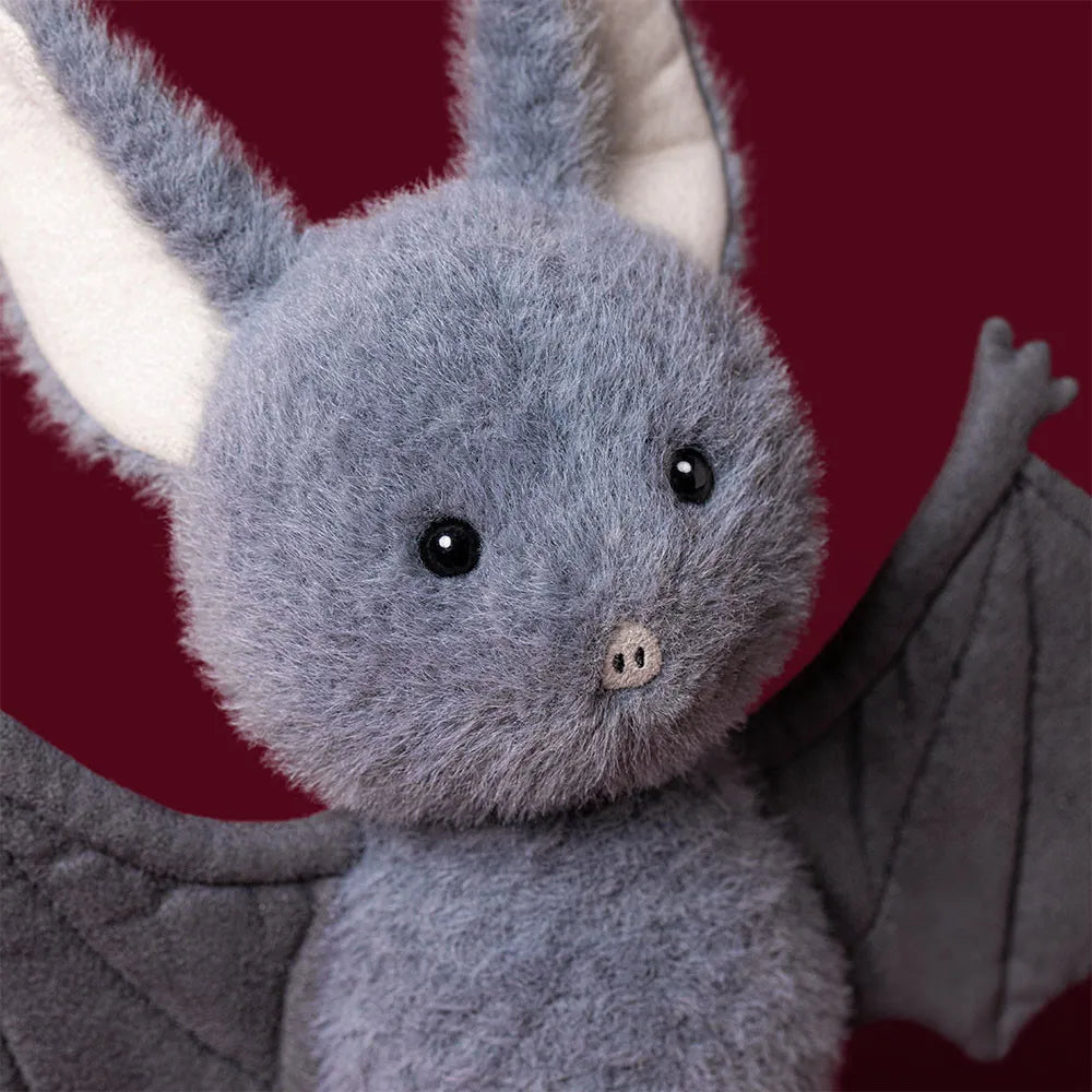 A plush bat toy with soft gray fur, large black eyes, a small pink nose, and folded gray wings, shown against a dark red background.