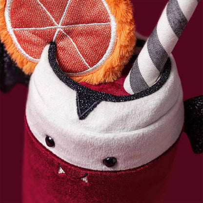 A close-up of a plush Halloween drink cup decoration, featuring a red velvet-like body, white plush whipped cream top, black glitter trim, a striped gray-and-white straw, and an orange slice accent on a dark red background.