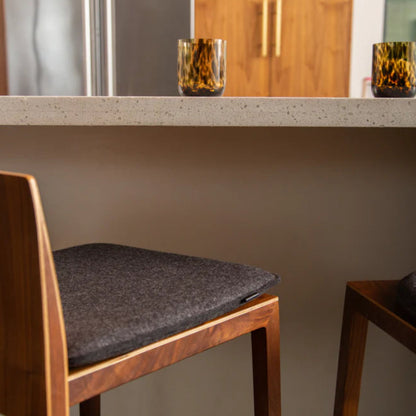 A charcoal-colored Merino wool square seat pad on a wooden chair in a modern kitchen setting, with a matching seat pad on a different chair in the background.