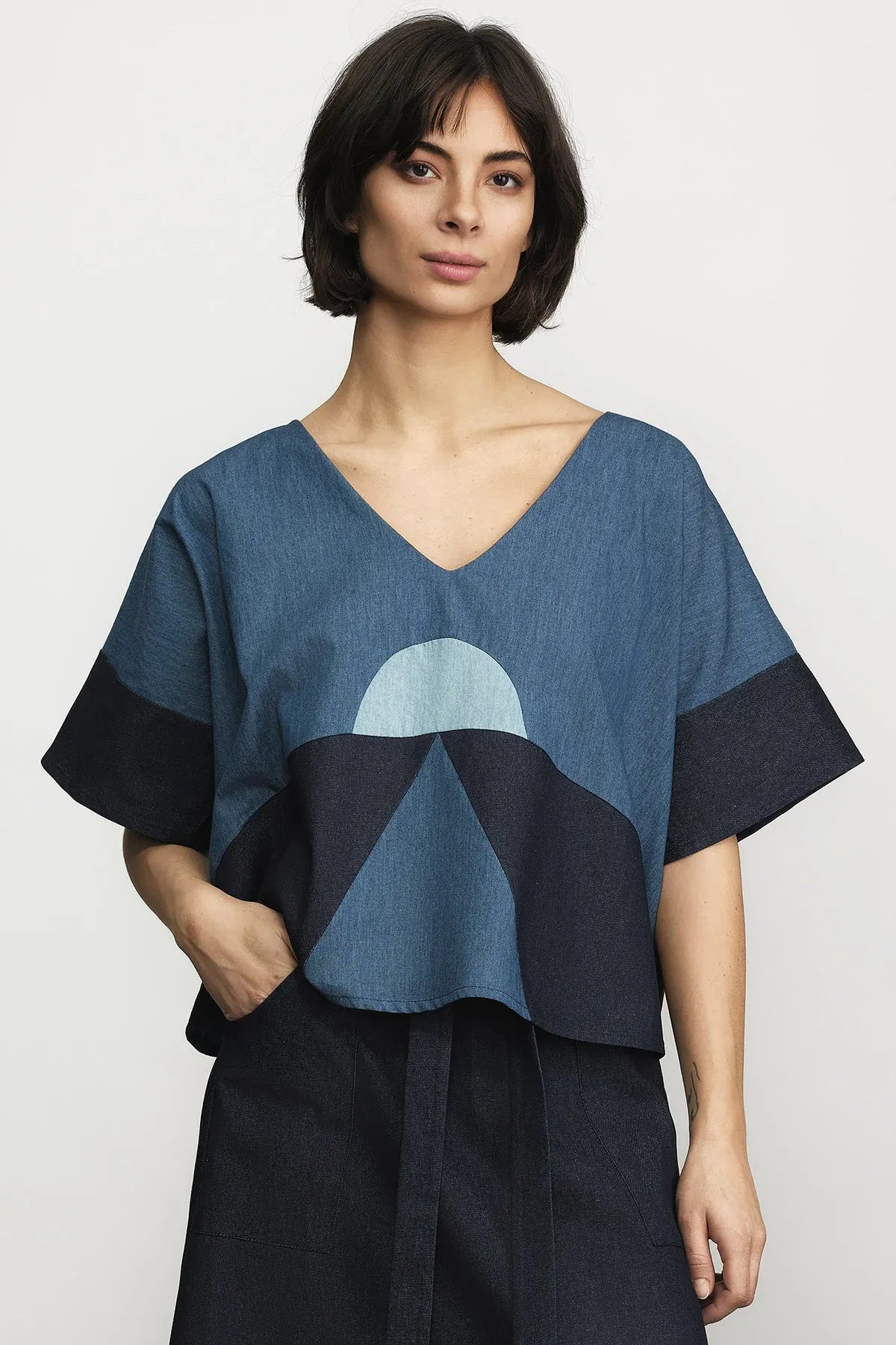 A woman modeling a loose, short-sleeve V-neck top with a geometric mountain pattern in shades of blue and navy, paired with dark navy bottoms, against a light gray studio background.