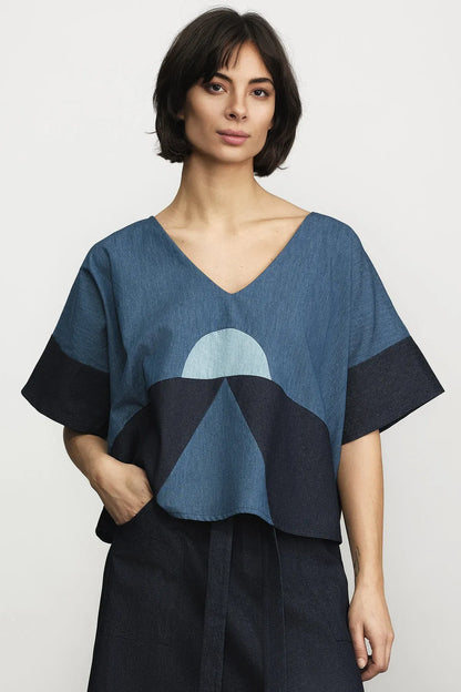 A woman modeling a loose, short-sleeve V-neck top with a geometric mountain pattern in shades of blue and navy, paired with dark navy bottoms, against a light gray studio background.