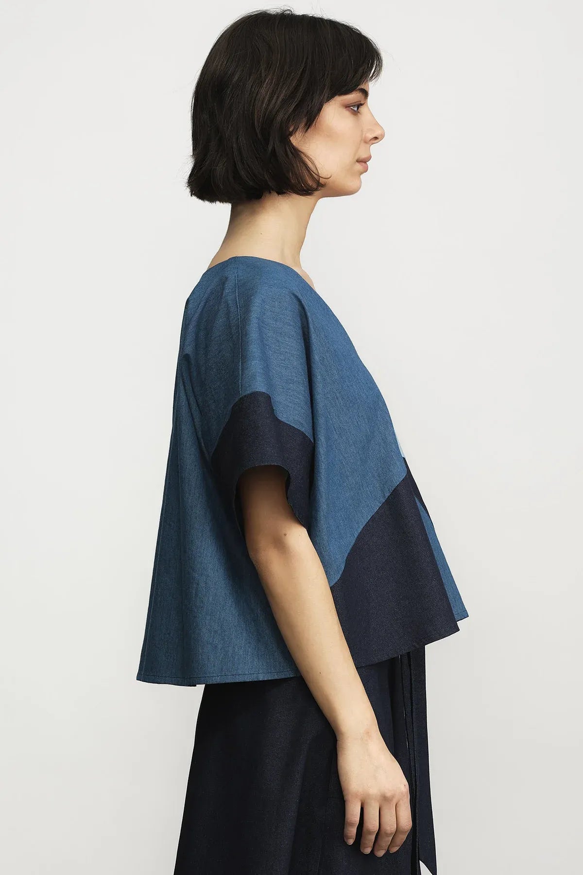 A woman shown in side profile wearing a color-blocked short-sleeve top in shades of blue and navy, paired with a dark skirt, against a plain light gray studio background.