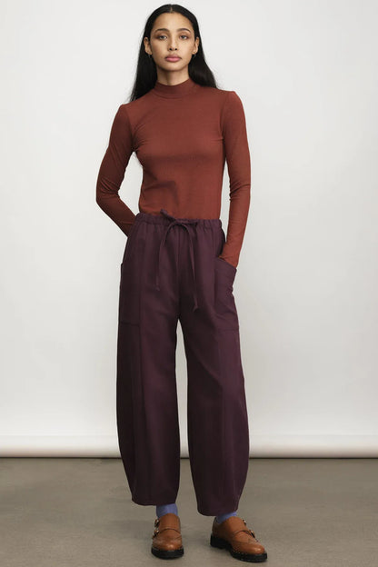 A woman modeling a rust-brown mock neck long-sleeve top and dark plum wide-leg drawstring pants, paired with brown leather loafers, against a neutral studio backdrop.