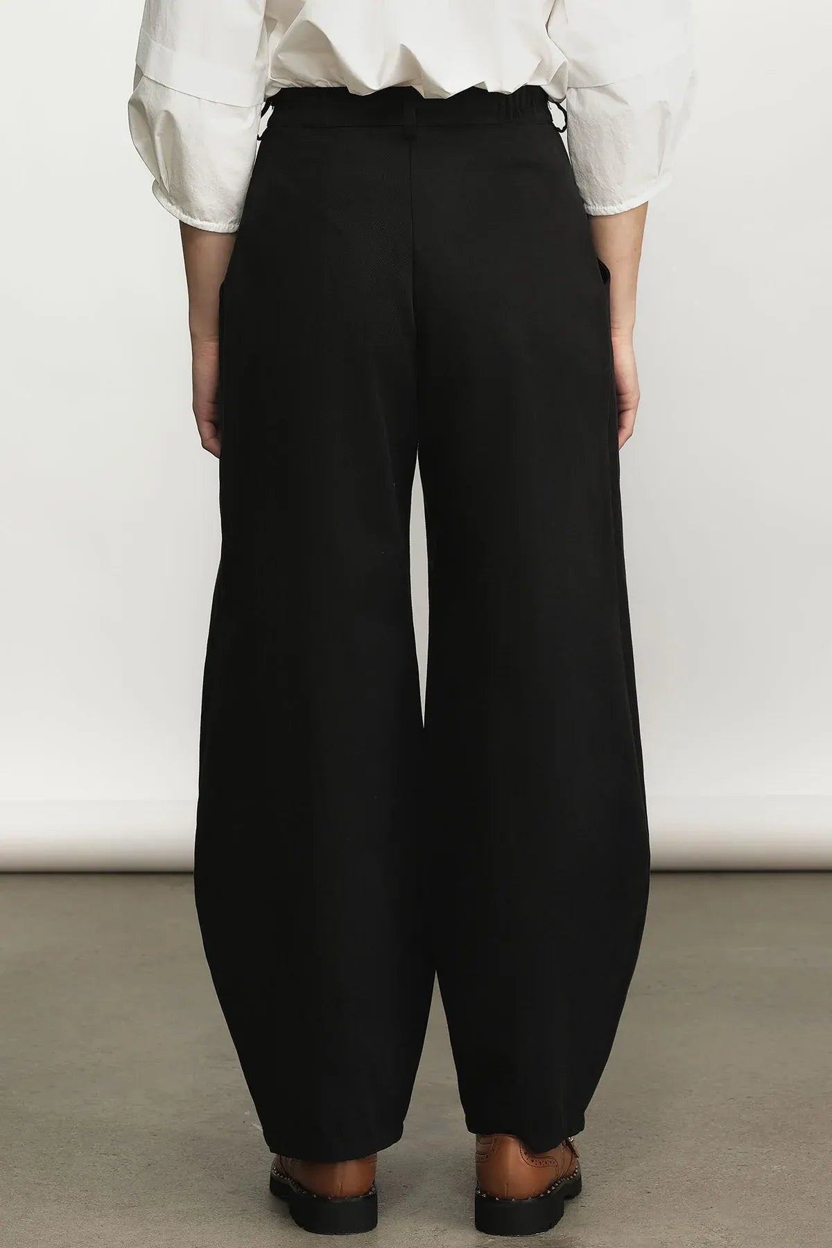 A person shown from the back wearing black wide-leg trousers with a white blouse and black shoes, standing against a plain light wall.