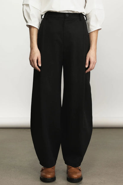 A person wearing loose black wide-leg trousers with a white long-sleeve top and brown shoes, standing against a plain light gray studio background.