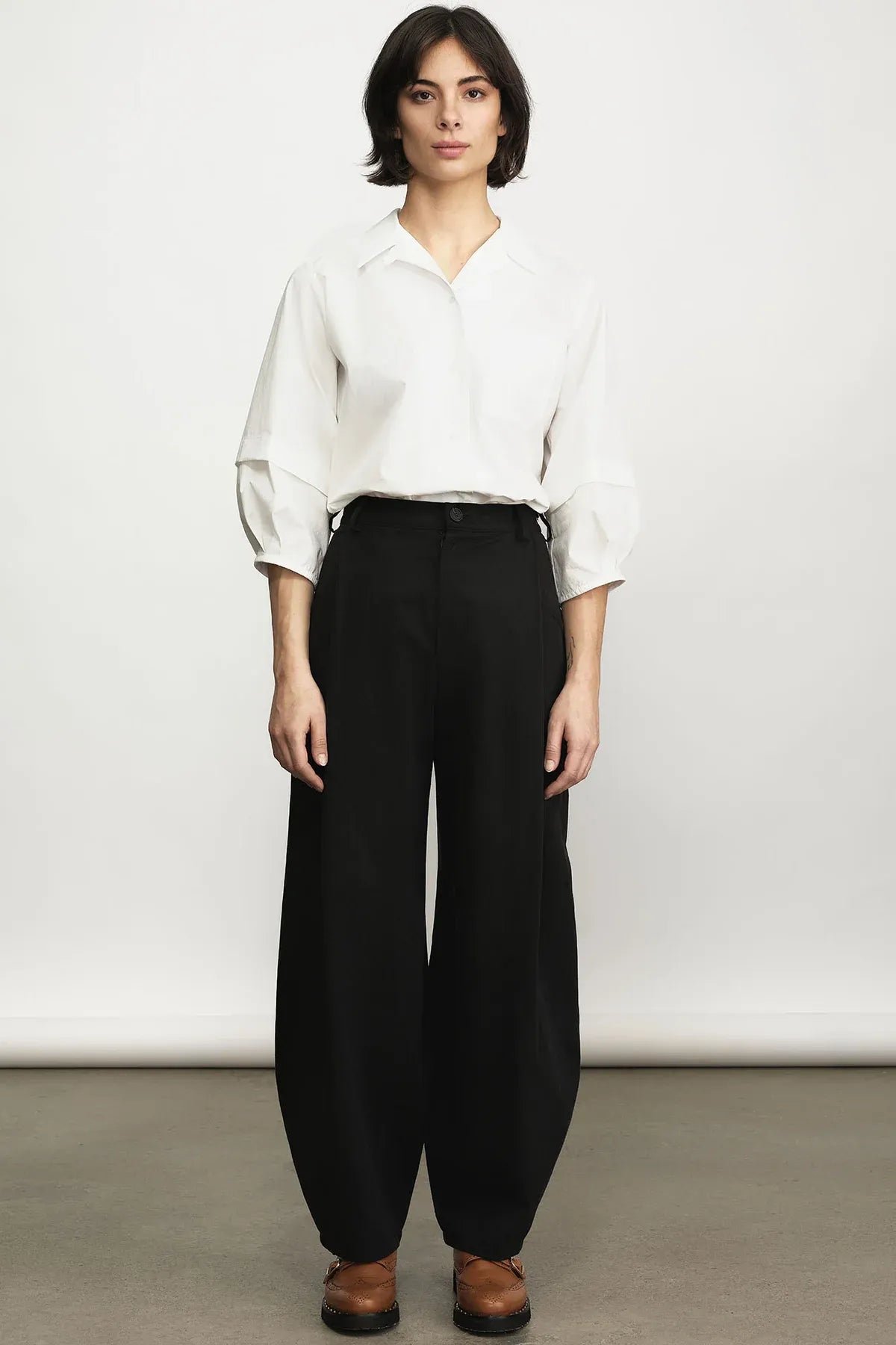 A model stands against a plain light gray studio wall wearing a white long-sleeve button-up shirt tucked into high-waisted black wide-leg trousers, with brown shoes.
