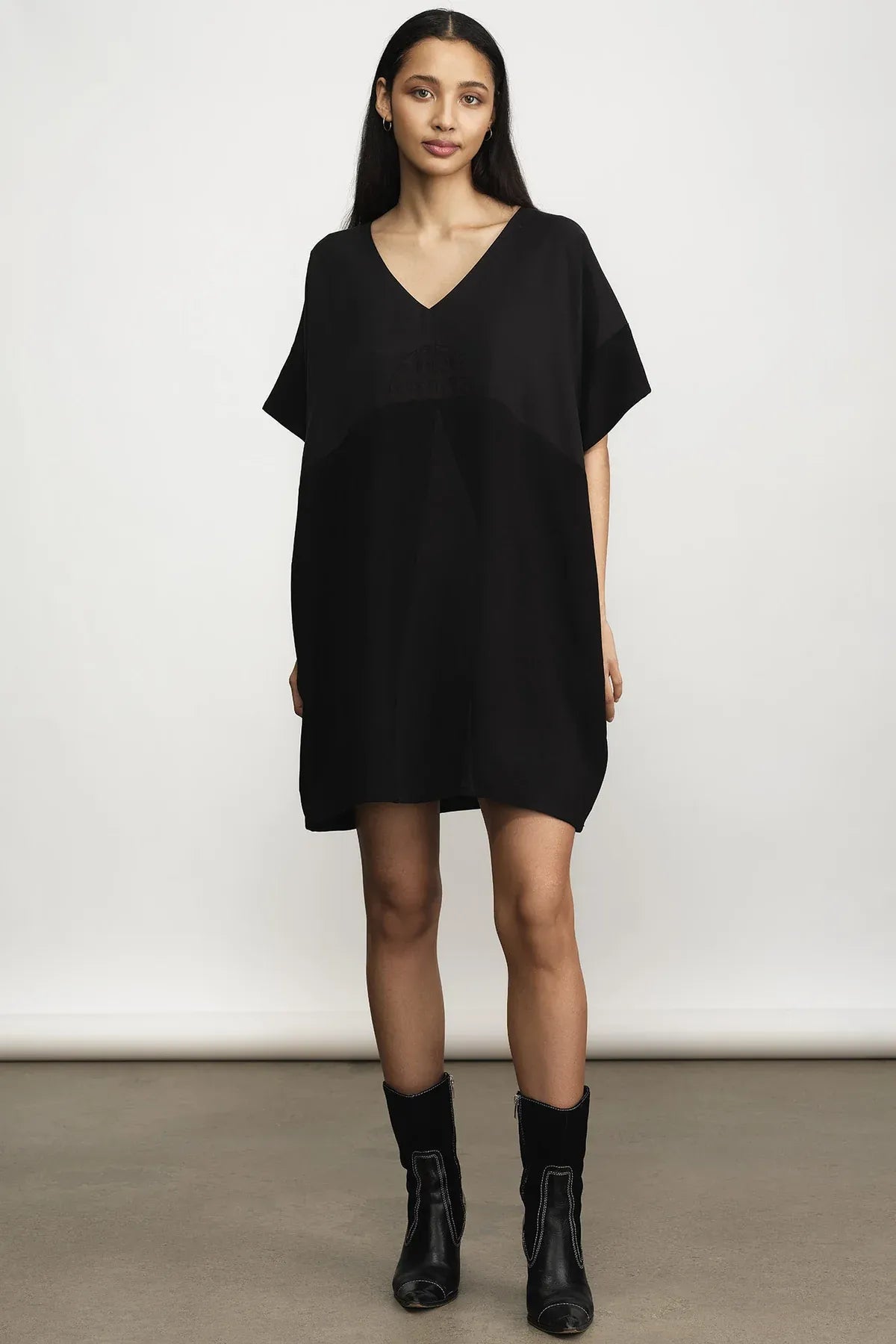 A woman modeling a loose-fitting black V-neck mini dress with short sleeves, paired with black ankle boots, against a plain light gray studio background.