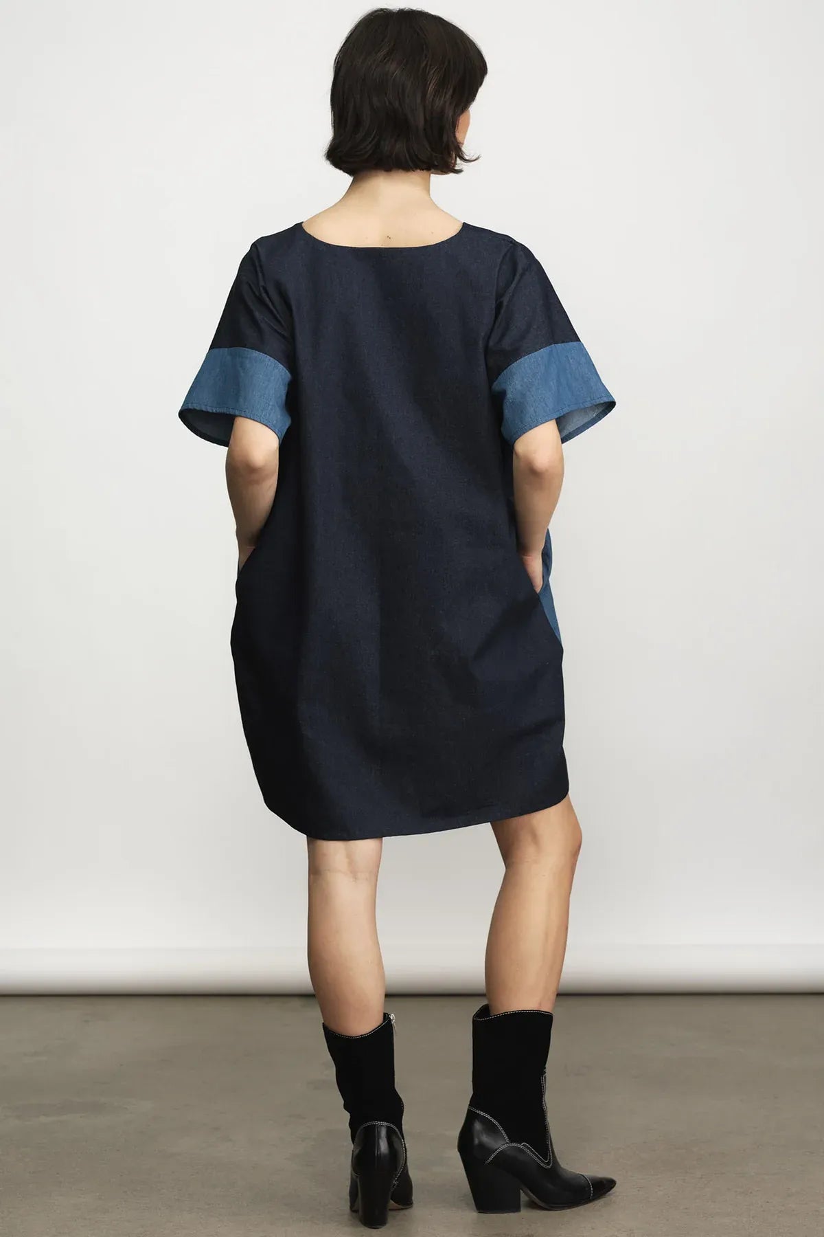 A model viewed from behind wearing a loose-fitting navy blue dress with contrasting medium blue sleeve cuffs, paired with black ankle boots, against a plain studio background.