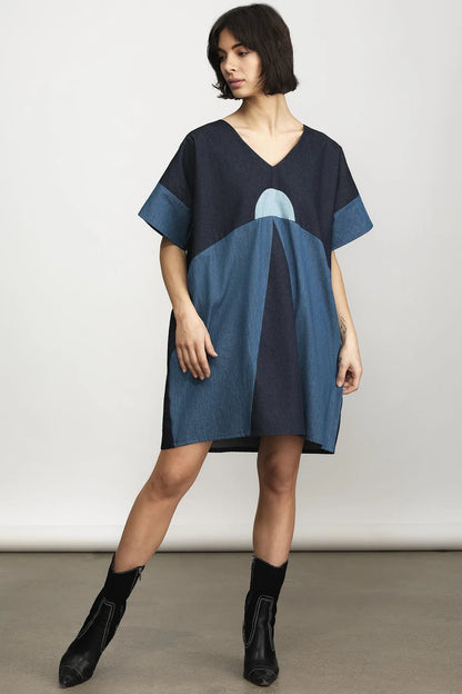 A woman models a loose, short-sleeve V-neck dress with a geometric color-block pattern in navy and medium blue, paired with black ankle boots, against a neutral studio background.