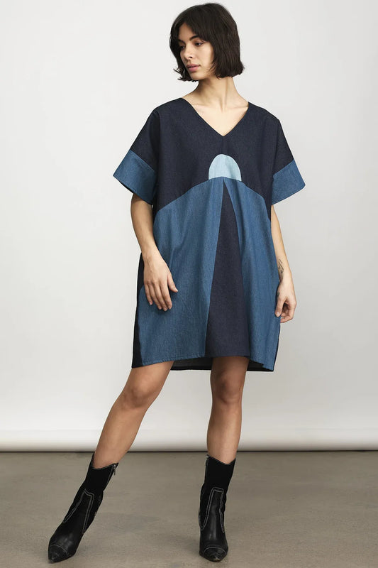 A woman models a loose, short-sleeve V-neck dress with a geometric color-block pattern in navy and medium blue, paired with black ankle boots, against a neutral studio background.