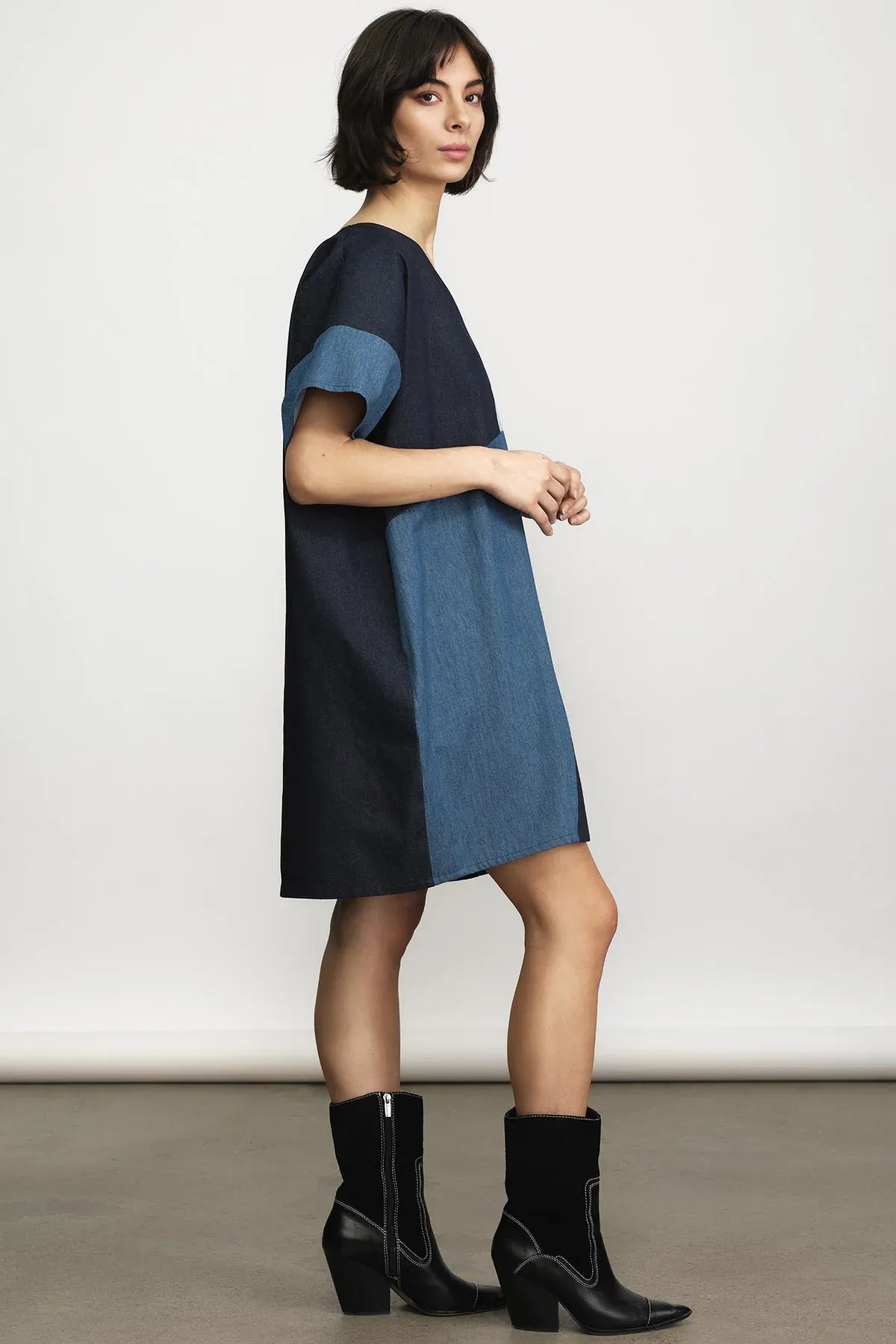 A woman modeling a color-block denim dress with a navy blue top and skirt, and medium blue short sleeves and side panels, paired with black ankle boots against a neutral studio background.