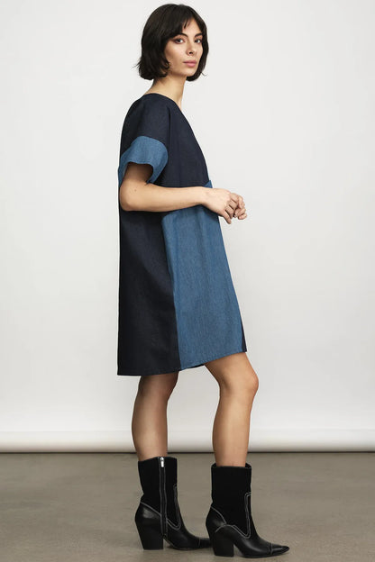A woman modeling a color-block denim dress with a navy blue top and skirt, and medium blue short sleeves and side panels, paired with black ankle boots against a neutral studio background.