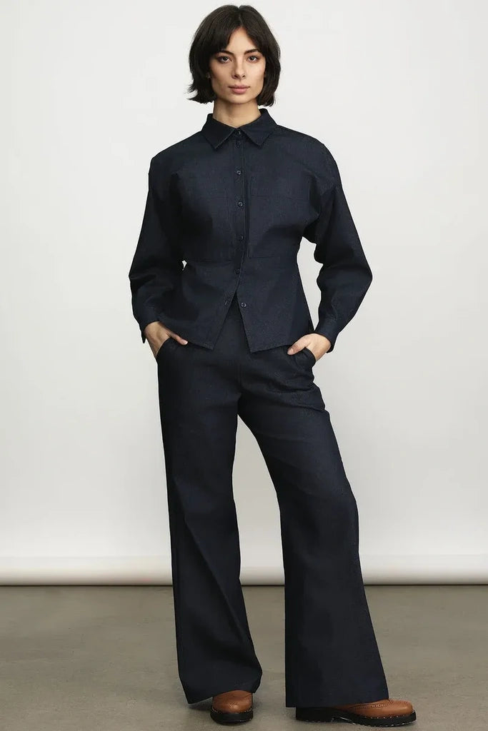 A model wearing a dark navy blue two-piece linen set consisting of a long-sleeve button-up shirt and matching wide-leg trousers, paired with brown leather shoes, standing against a neutral studio backdrop.