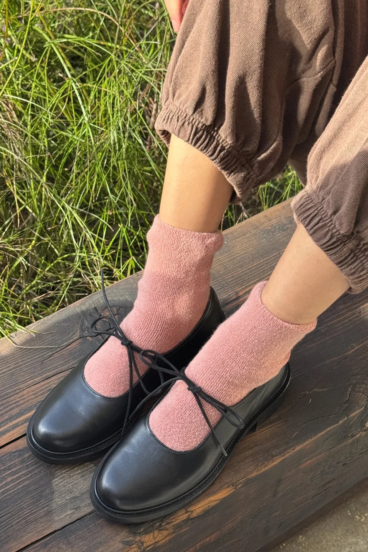 Person wearing black shoes and pink socks sitting on a wooden bench with grass in the background